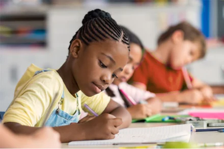 Crianças sentadas escrevendo no caderno, em primeiro foco, há uma garotinha de macação e camiseta amarela